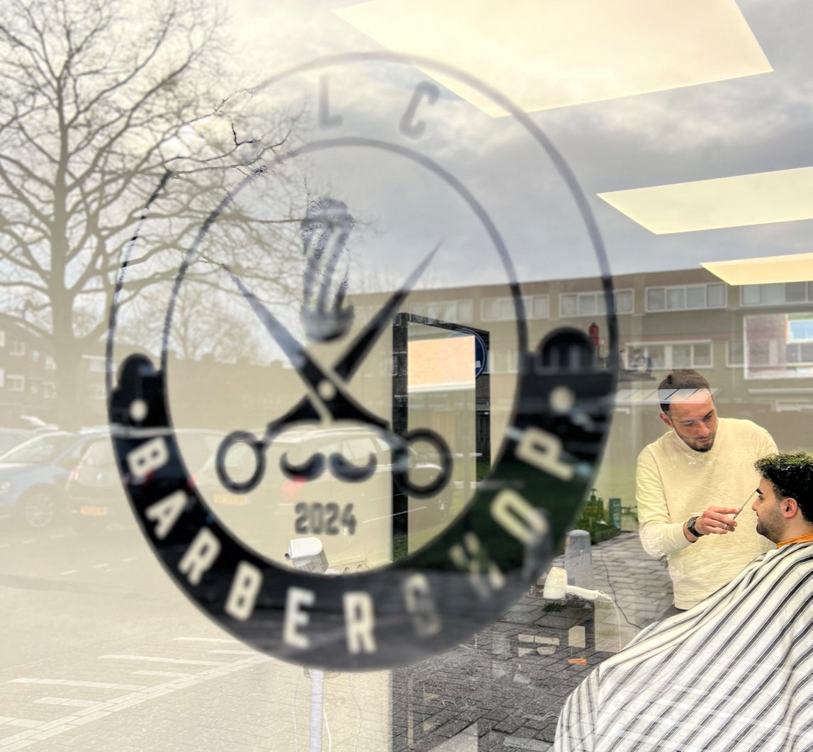 Ercan knipt een klant, gezien door het raam van de zaak met het SLC Barbershop logo op het glas
