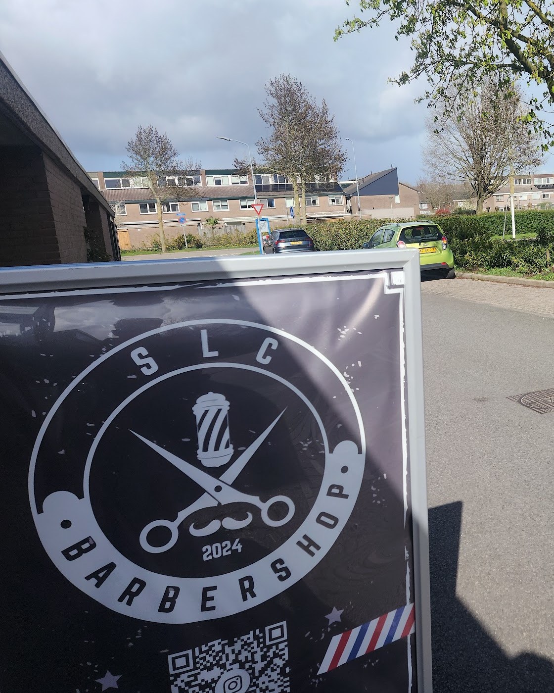 Het reclamebord van SLC Barbershop met het ronde logo op de stoep voor de zaak